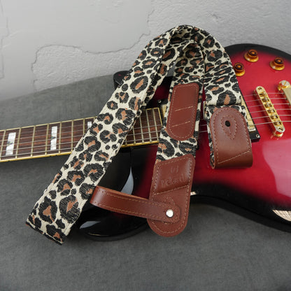 A leopard print guitar strap with brown leather accents, draped over a red electric guitar on a gray surface against a light gray wall.