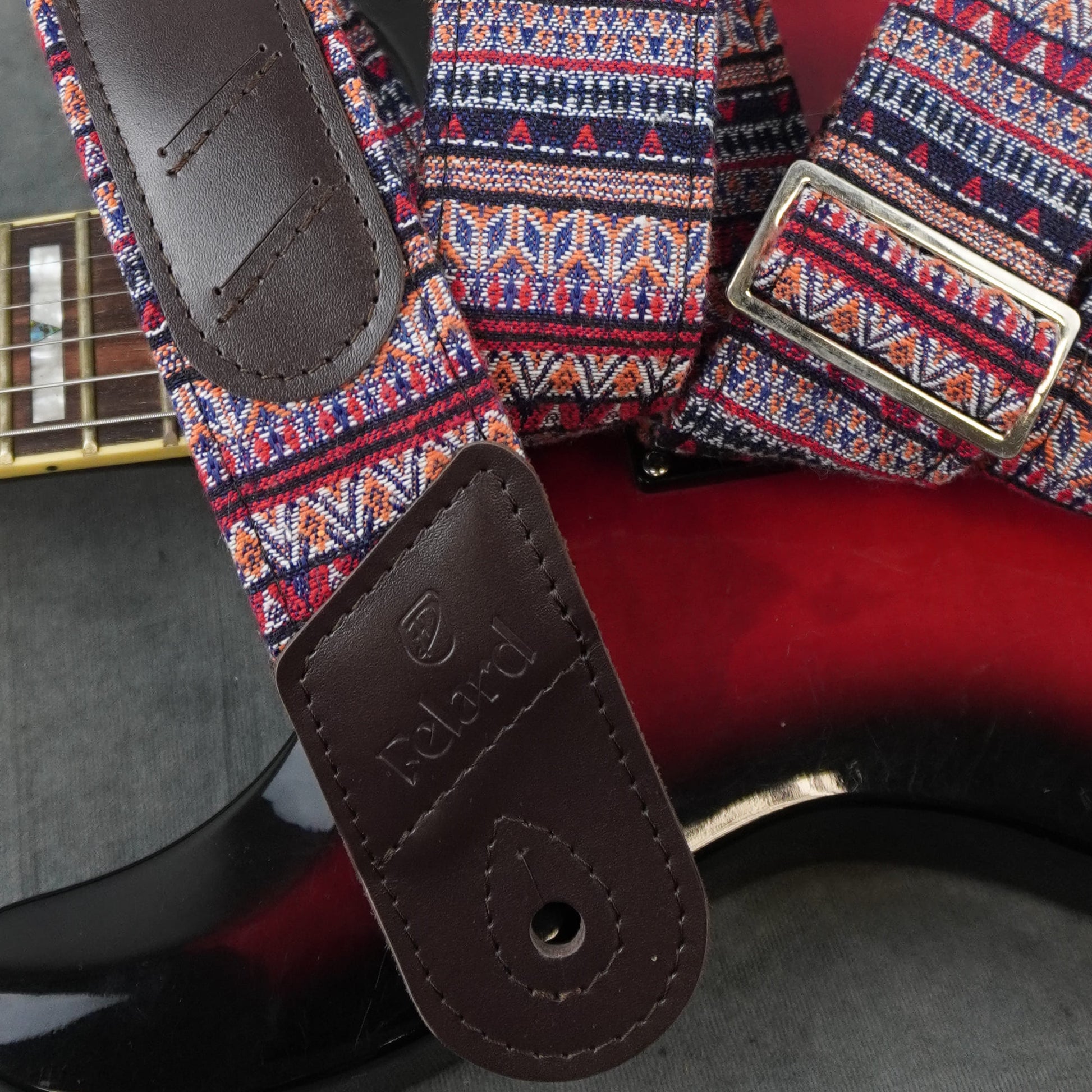 A close-up of a guitar strap with a colorful geometric pattern in red, blue, orange, and white, featuring dark brown leather end pieces and a silver metal buckle. The strap is draped over a red and black electric guitar.