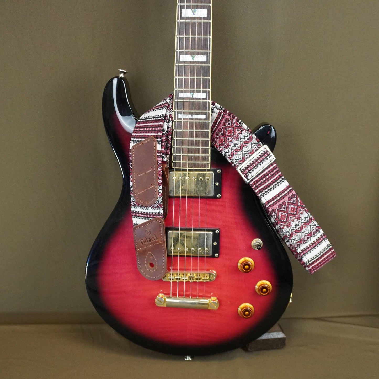  red, white, and black patterned guitar strap and a matching brown pickguard, displayed against a solid brown background.