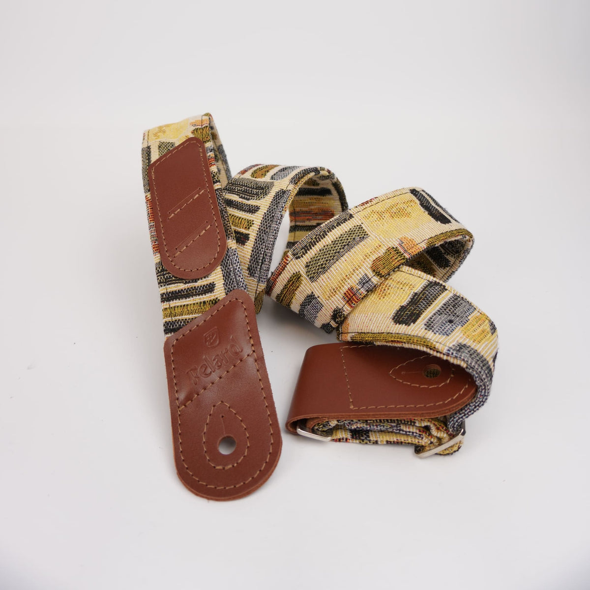 Guitar strap with a multicolor geometric pattern in beige, brown, and black, featuring brown leather end caps and a buckle closure.