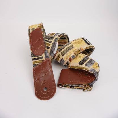 Guitar strap with a multicolor geometric pattern in beige, brown, and black, featuring brown leather end caps and a buckle closure.