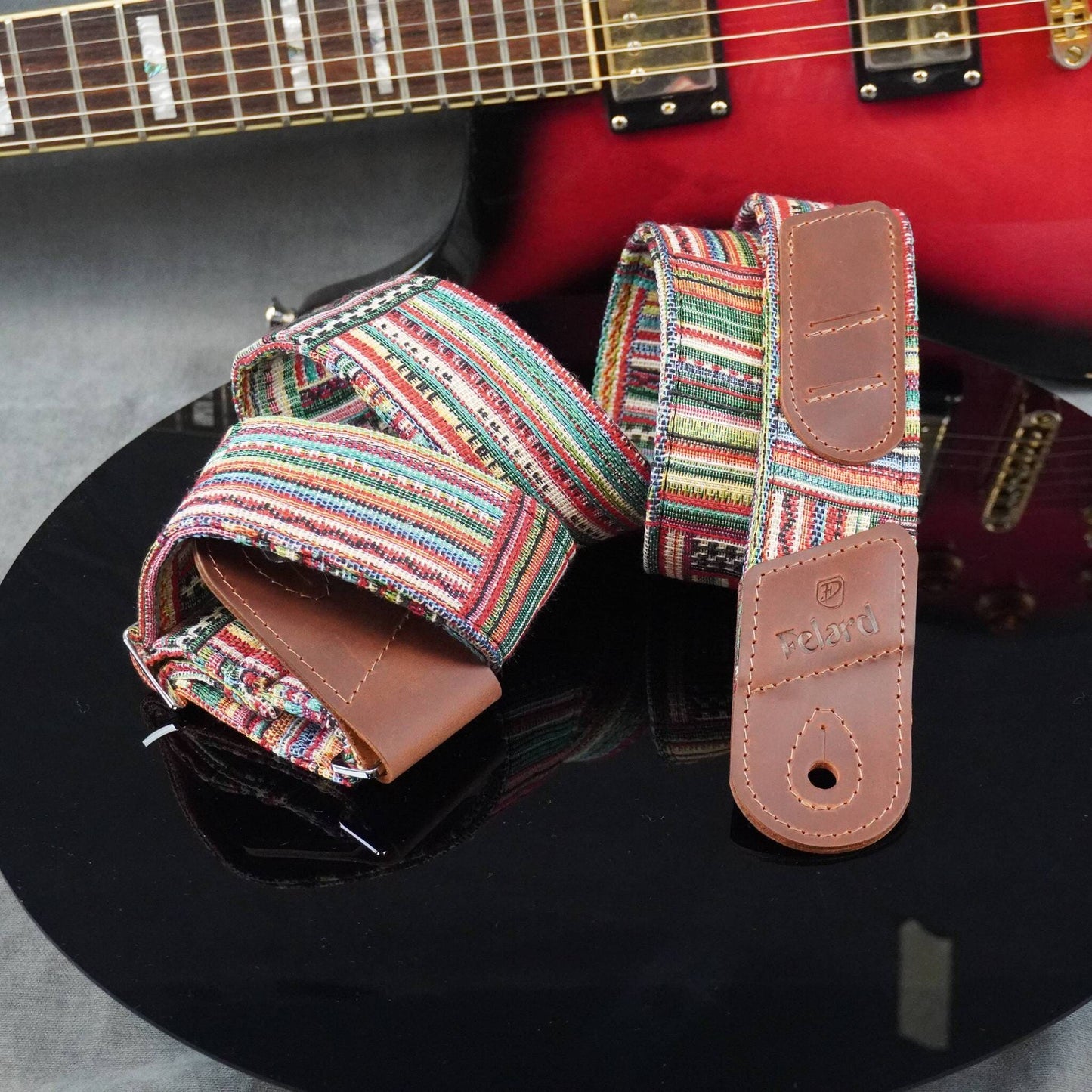 A guitar strap with a colorful striped woven pattern and brown leather ends, displayed on a glossy black surface with a red electric guitar in the background.