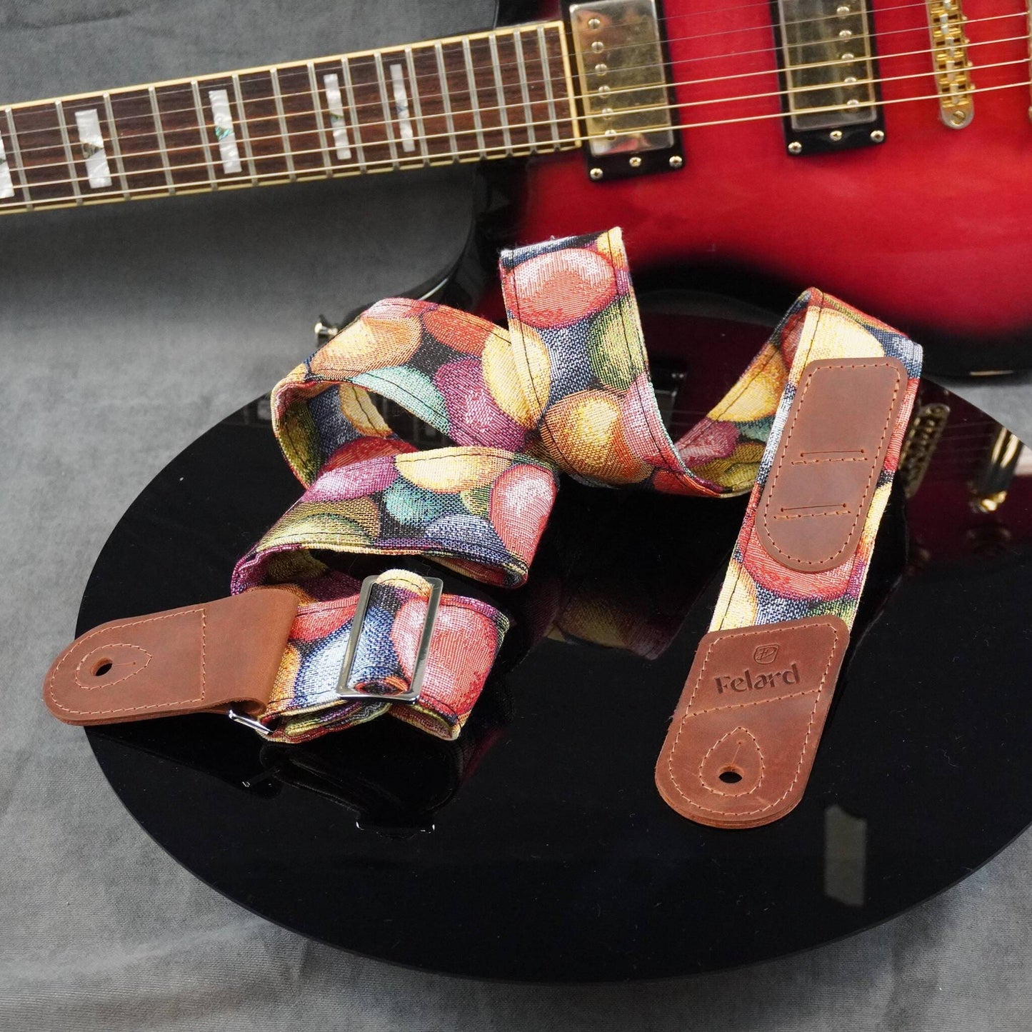 A guitar strap with a colorful fruit print and brown leather ends, displayed on a black surface with a red electric guitar in the background.
