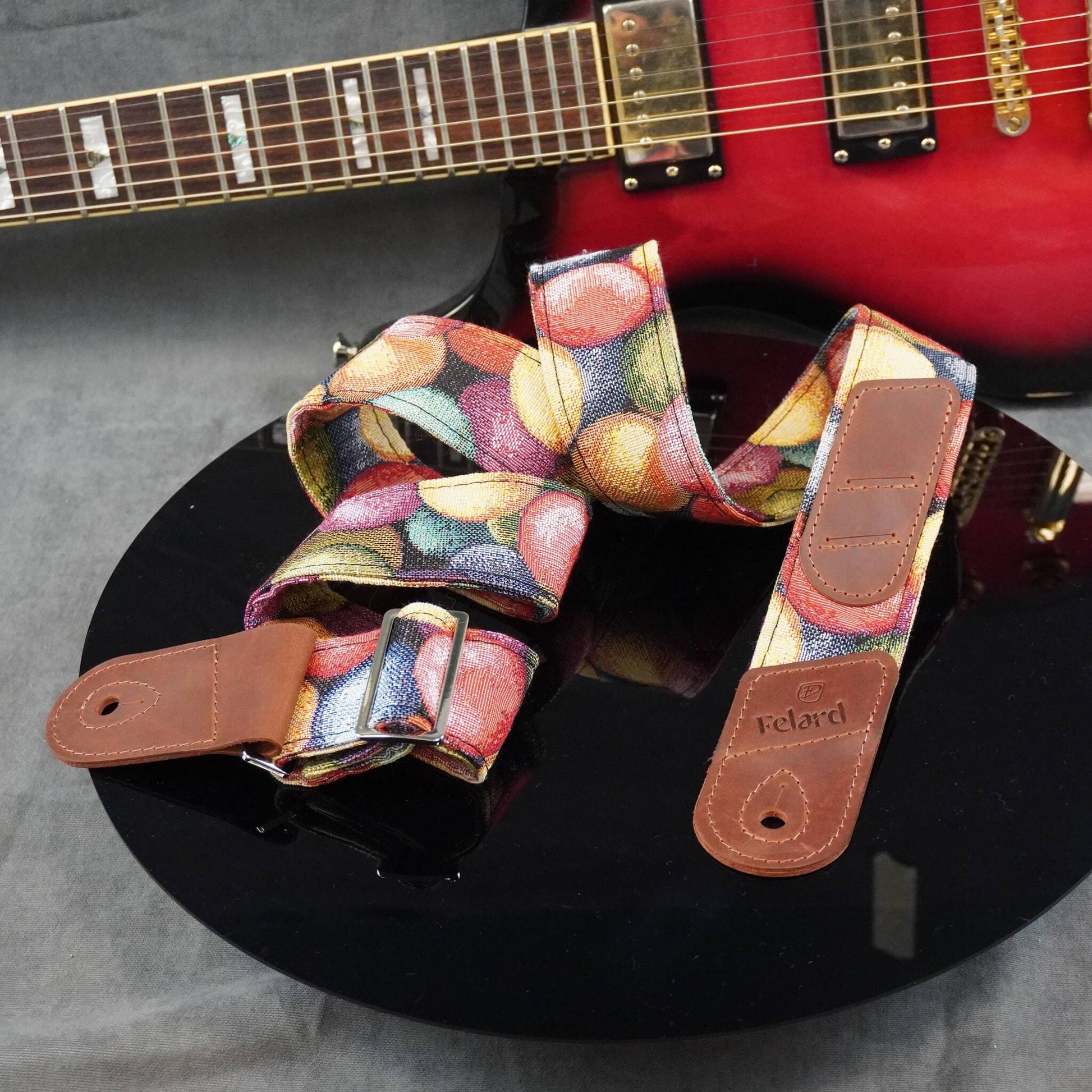 A guitar strap with a colorful fruit print and brown leather ends, displayed on a black surface with a red electric guitar in the background.
