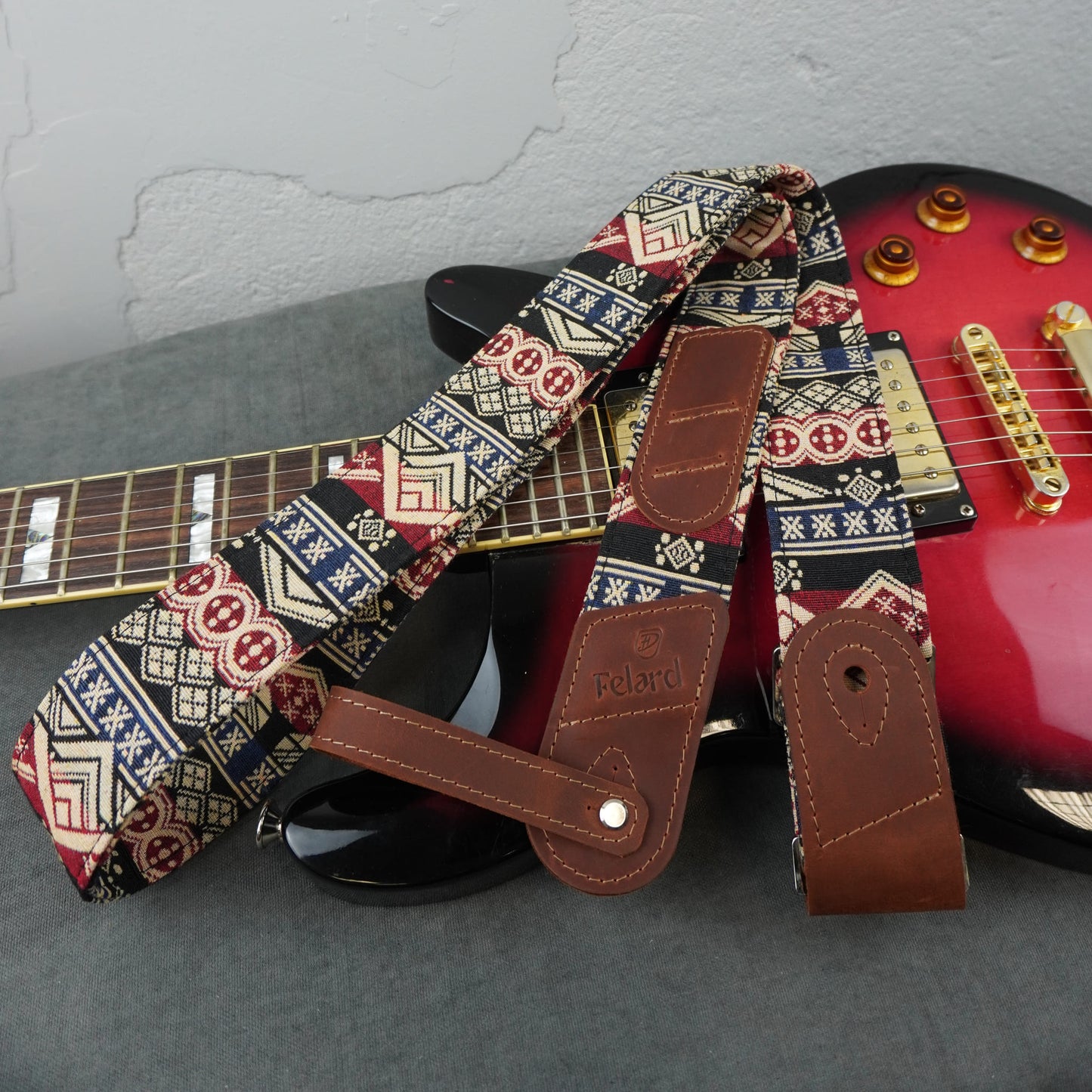 A guitar strap with a brown leather end and a geometric patterned fabric body, laid across a red electric guitar on a gray surface against a light gray wall.