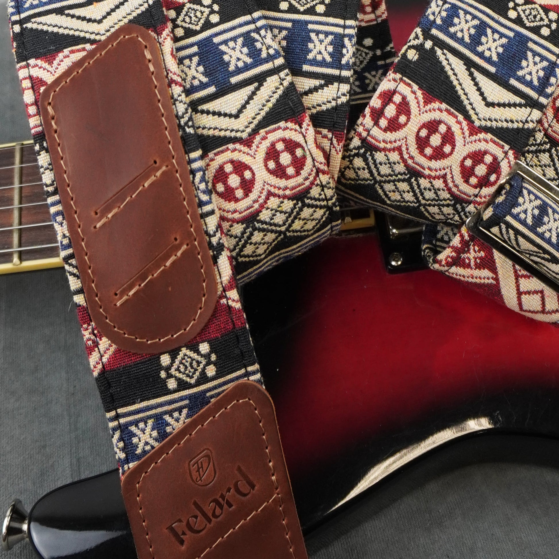 A close-up of a guitar strap with a brown leather end piece and a patterned woven fabric body featuring red, blue, and cream geometric motifs, shown attached to an electric guitar.