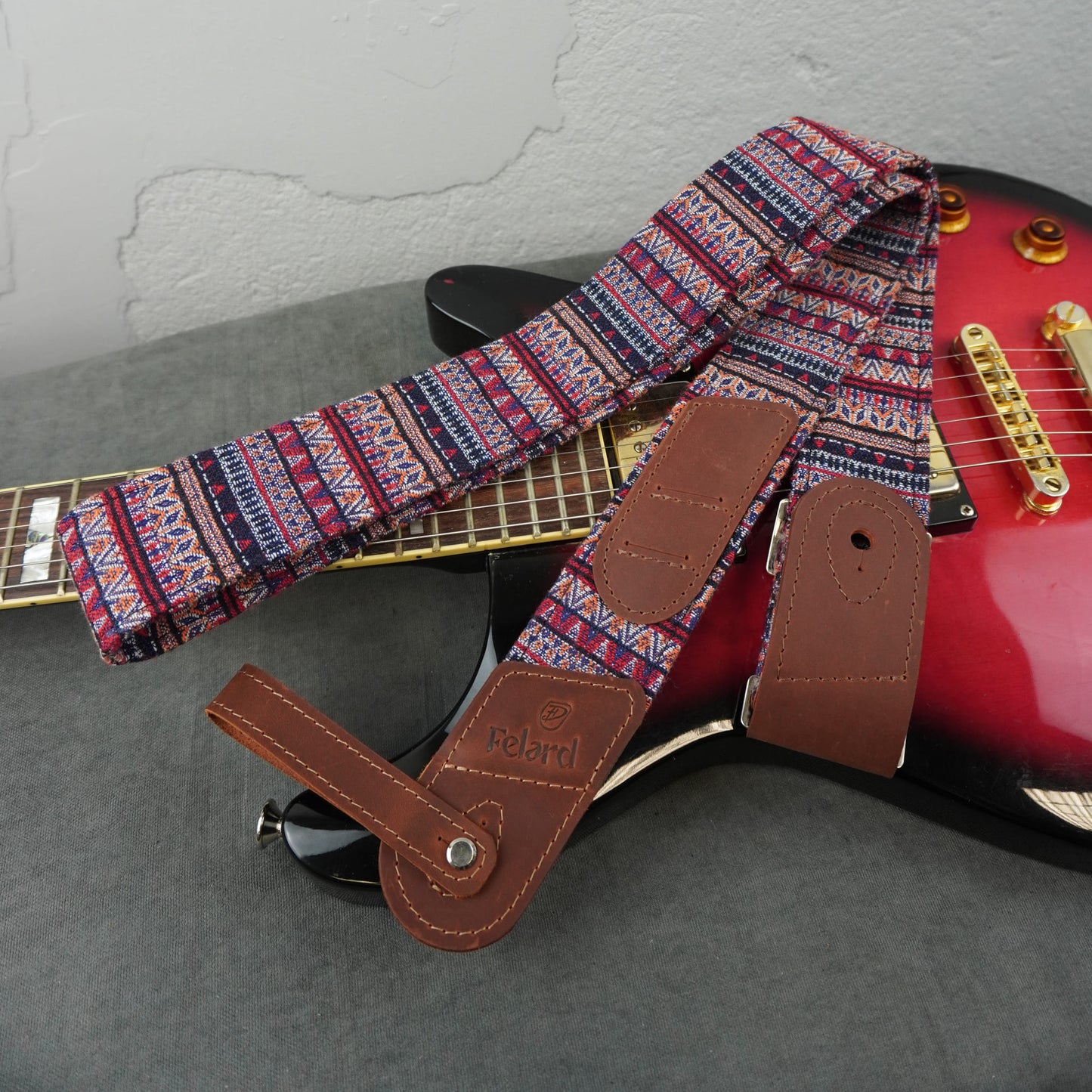 guitar strap with a colorful geometric patterned fabric and brown leather attachments, shown resting across a red electric guitar on a gray surface.