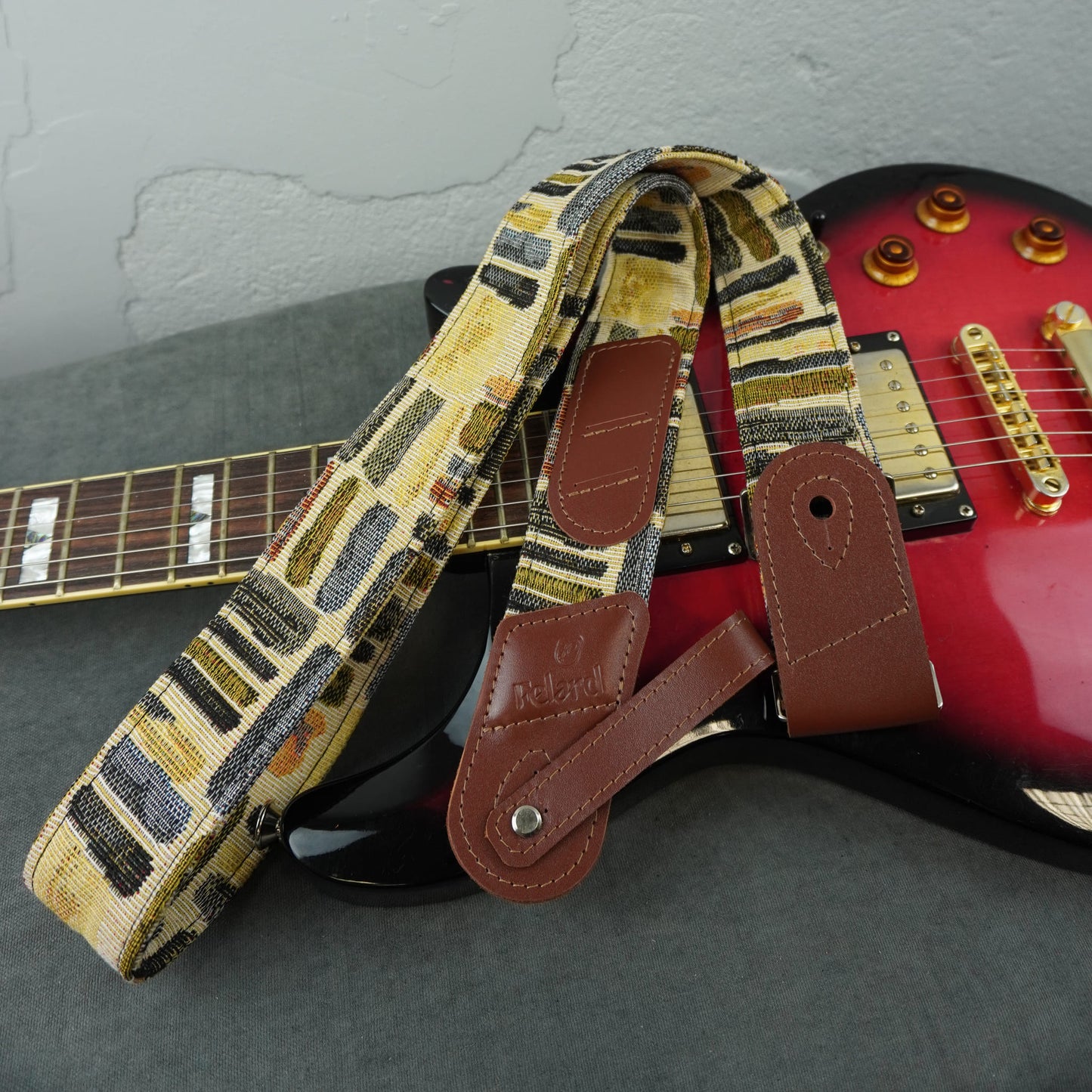 guitar strap with a beige, brown, and black geometric pattern and brown leather ends, draped over a red electric guitar on a gray surface.