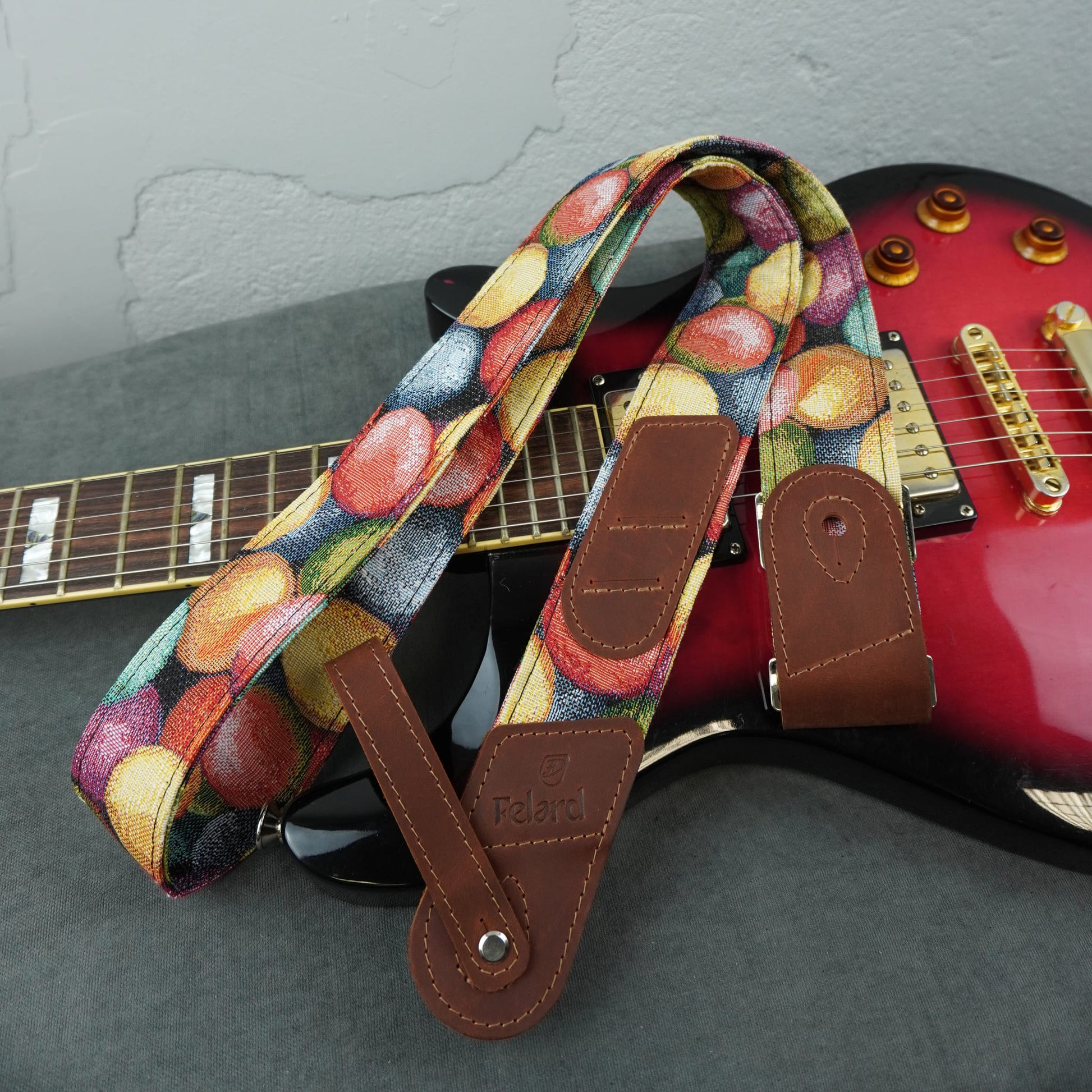A guitar strap with a colorful fruit print and brown leather ends, draped over a red electric guitar on a gray surface.