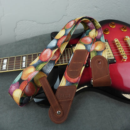 A guitar strap with a colorful fruit print and brown leather ends, draped over a red electric guitar on a gray surface.