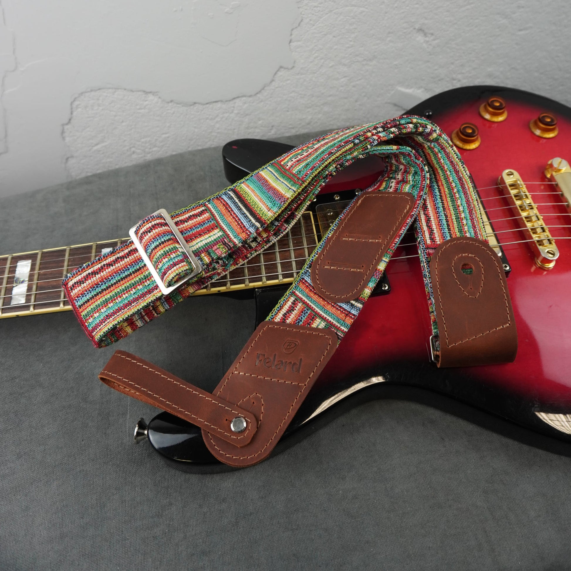 A guitar strap with a colorful striped woven fabric body and brown leather ends, shown draped over a red electric guitar on a gray surface against a light gray wall.