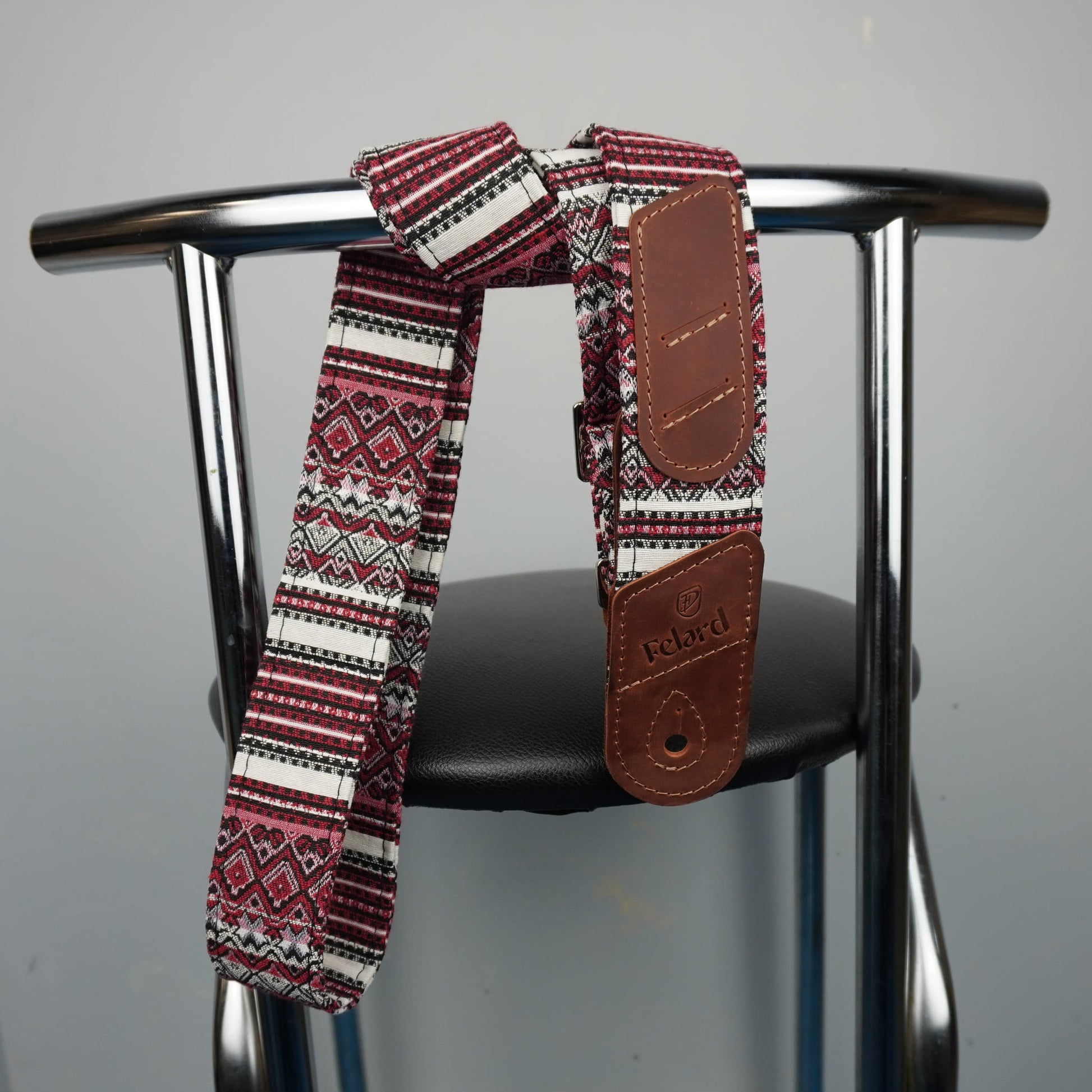 A patterned fabric strap with brown leather patches, draped over the back of a metal chair with a black seat against a plain gray wall.