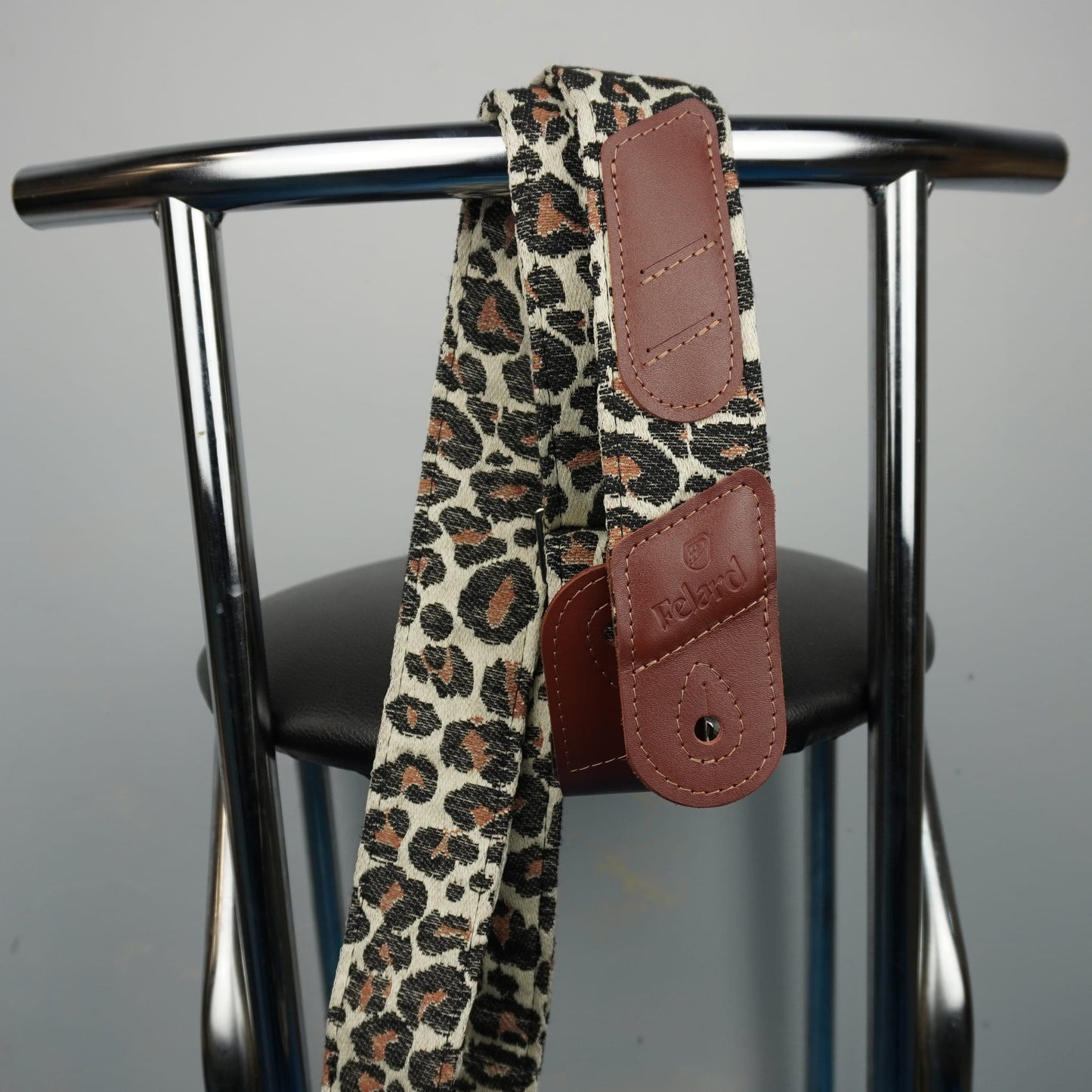 leopard print guitar strap with brown faux-leather end pieces, draped over the back of a black chair with a chrome metal frame.