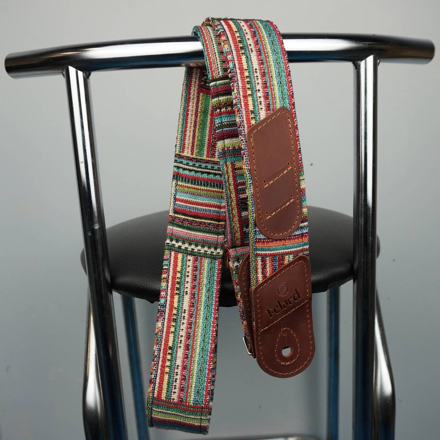 A multicolor guitar strap with brown leather patches hangs from the backrest of a metal chair against a plain gray background.