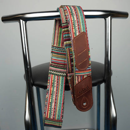 A multicolor guitar strap with brown leather patches hangs from the backrest of a metal chair against a plain gray background.