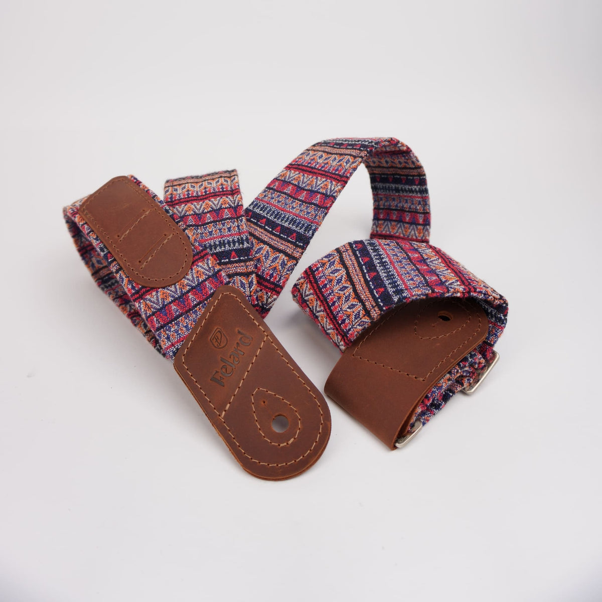A woven guitar strap with a multicolor geometric pattern in red, blue, and pink, featuring brown leather end caps and a silver metal buckle, displayed on a clean white background.