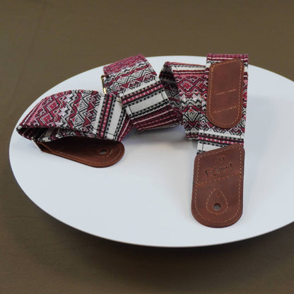 Guitar strap with a red, white, and black geometric pattern and brown leather ends, displayed on a white plate against a brown background.