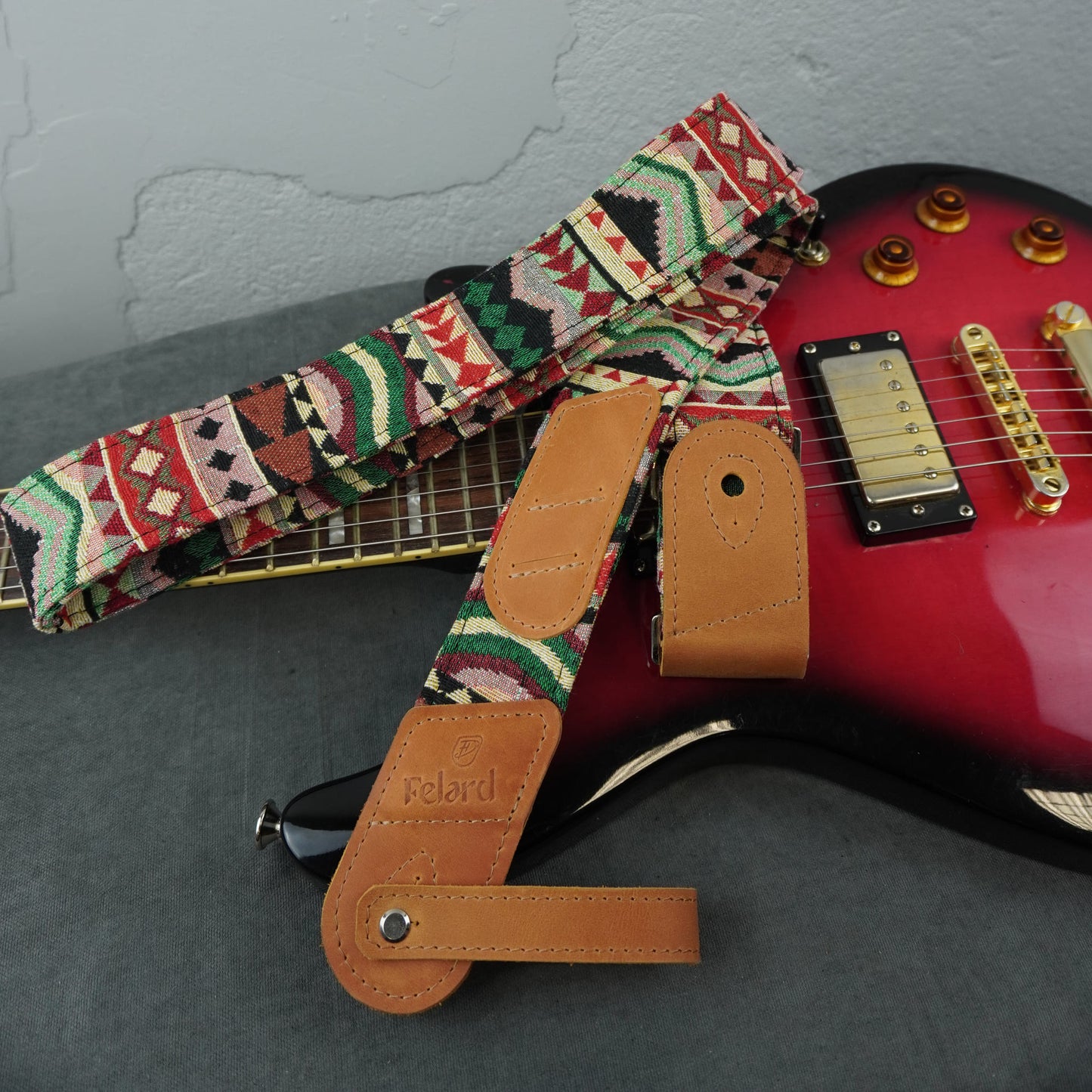 A guitar strap with a colorful geometric pattern and tan leather ends is draped over a red electric guitar on a gray surface against a light gray wall.