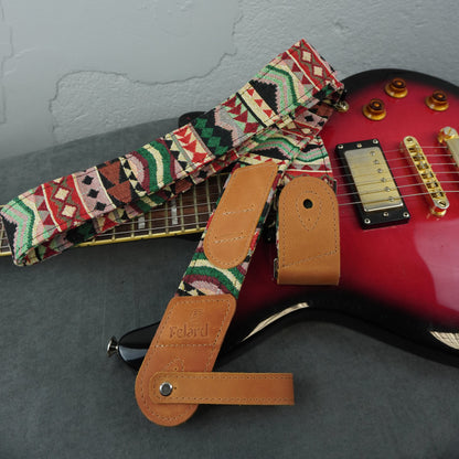 A guitar strap with a colorful geometric pattern and tan leather ends is draped over a red electric guitar on a gray surface against a light gray wall.