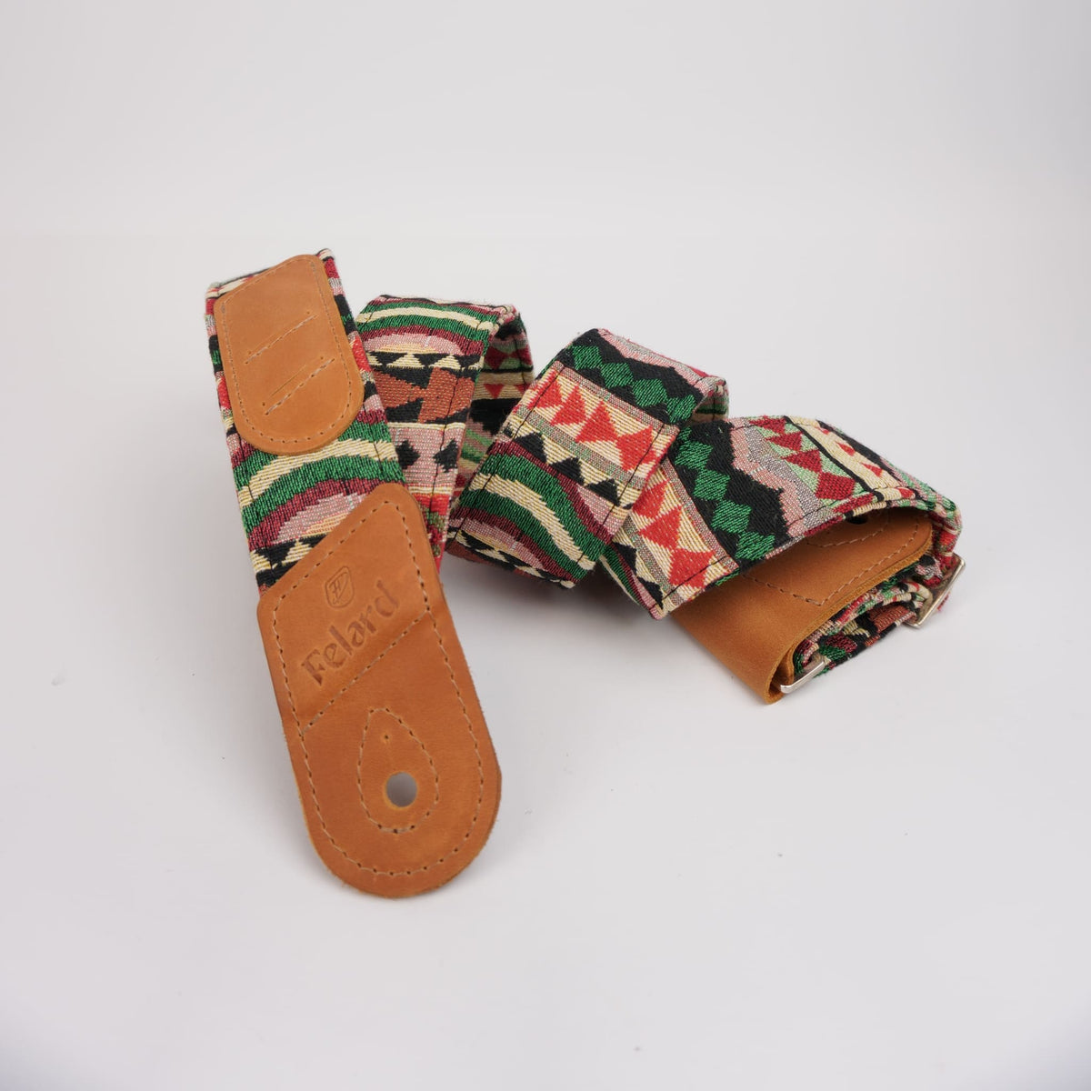 A woven fabric guitar strap with a Southwestern geometric kilim pattern in red, green, beige, and black, featuring tan leather ends.