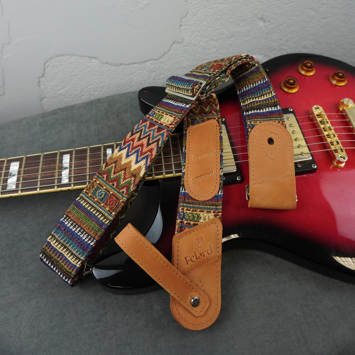 A colorful patterned electric guitar strap with tan leather end pieces rests across two guitars on a gray surface against a light gray wall.