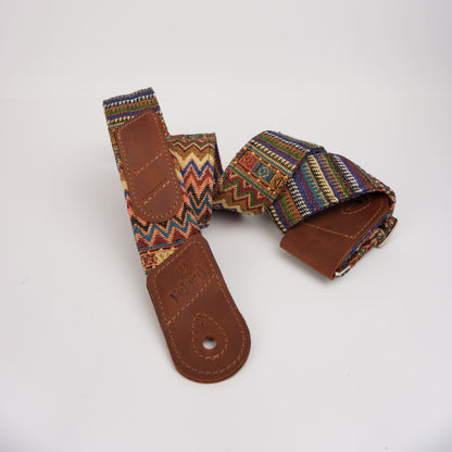 Guitar strap featuring a brown leather loop and a colorful woven fabric strap with geometric patterns in red, blue, yellow, and green, displayed on a white background.
