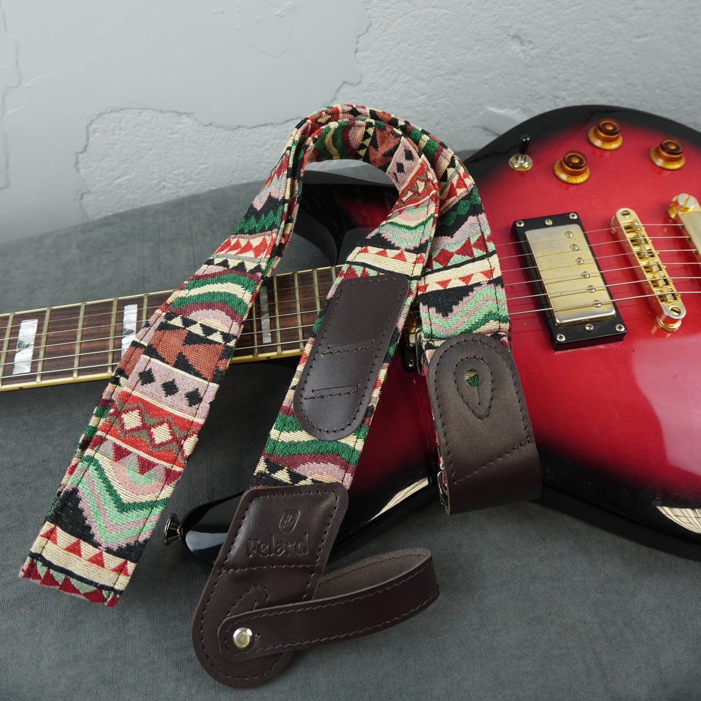 A patterned electric guitar strap with dark brown leather ends and a geometric Southwestern-style print, shown draped over a red electric guitar on a gray surface.