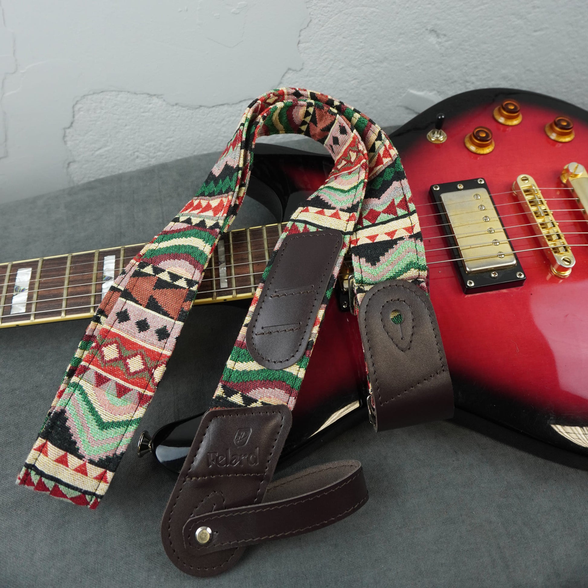 A patterned electric guitar strap with dark brown leather ends and a geometric Southwestern-style print, shown draped over a red electric guitar on a gray surface.