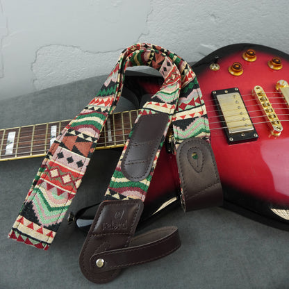 A patterned electric guitar strap with dark brown leather ends and a geometric Southwestern-style print, shown draped over a red electric guitar on a gray surface.
