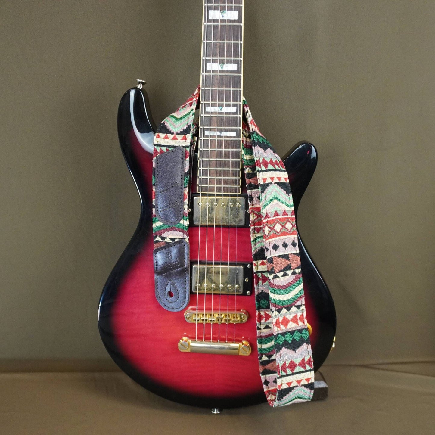 A red and black electric guitar with a colorful geometric patterned strap, photographed against a plain brown backdrop.