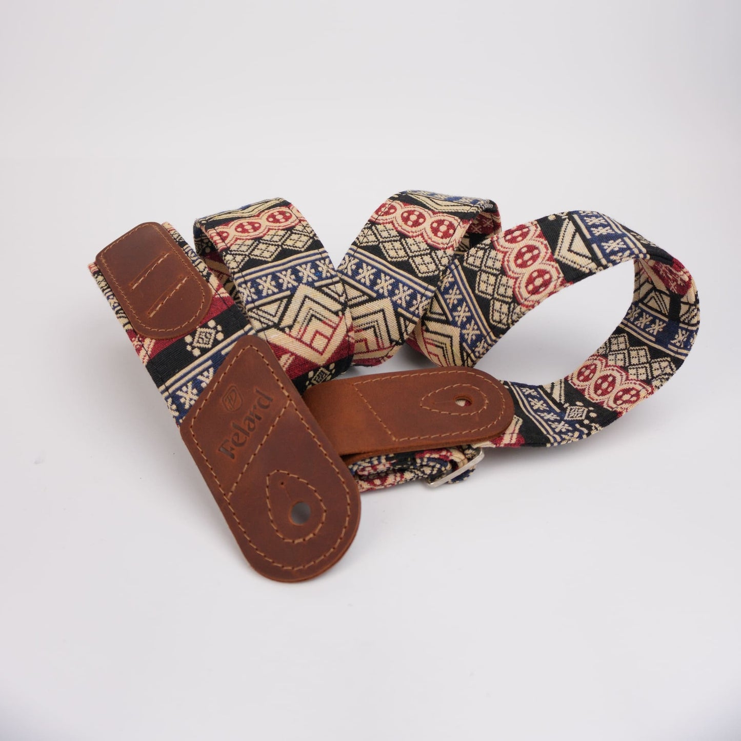 A fabric guitar strap with a multicolor geometric pattern in beige, navy, red, and black, featuring brown leather hardware and a visible 'Felard' brand stamp.