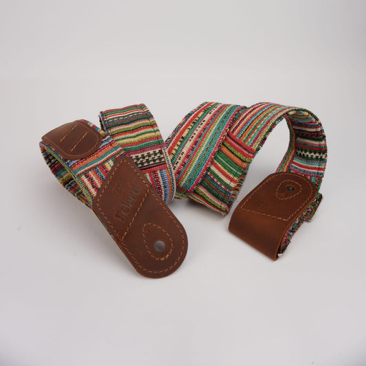 Guitar strap featuring a colorful woven textile with red, green, yellow, black, and white stripes, finished with brown leather ends and a metal snap button.