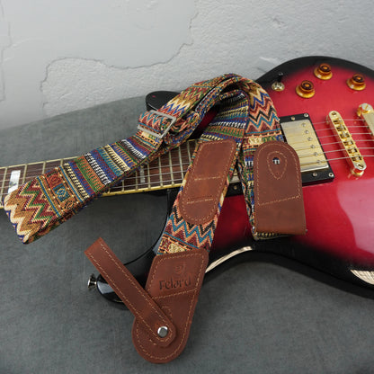 A guitar strap with a brown leather end pieces and a colorful geometric patterned woven fabric body, laid over a red electric guitar on a gray surface.
