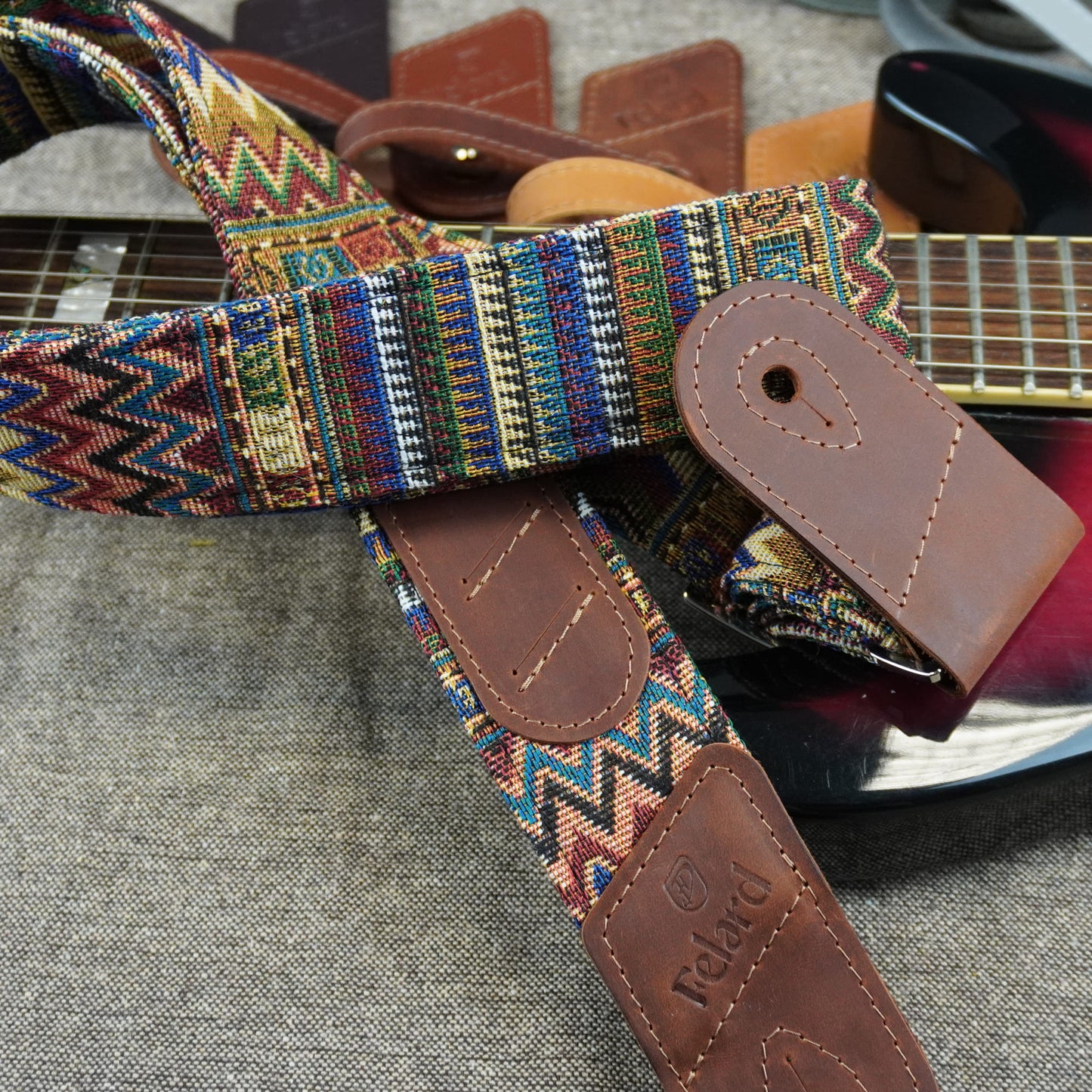 guitar strap with a colorful geometric woven pattern and brown leather end pieces, draped over a guitar on a gray fabric surface.