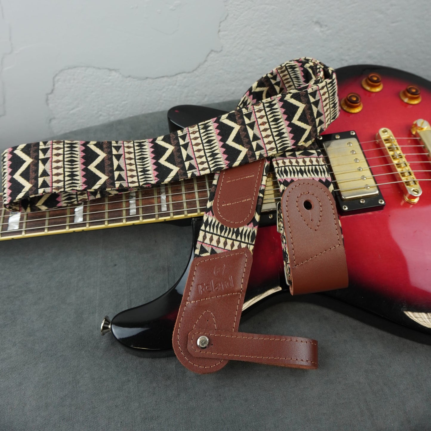 showing a patterned guitar strap with brown leather ends. The strap has a black, beige, and pink geometric tribal-style print.