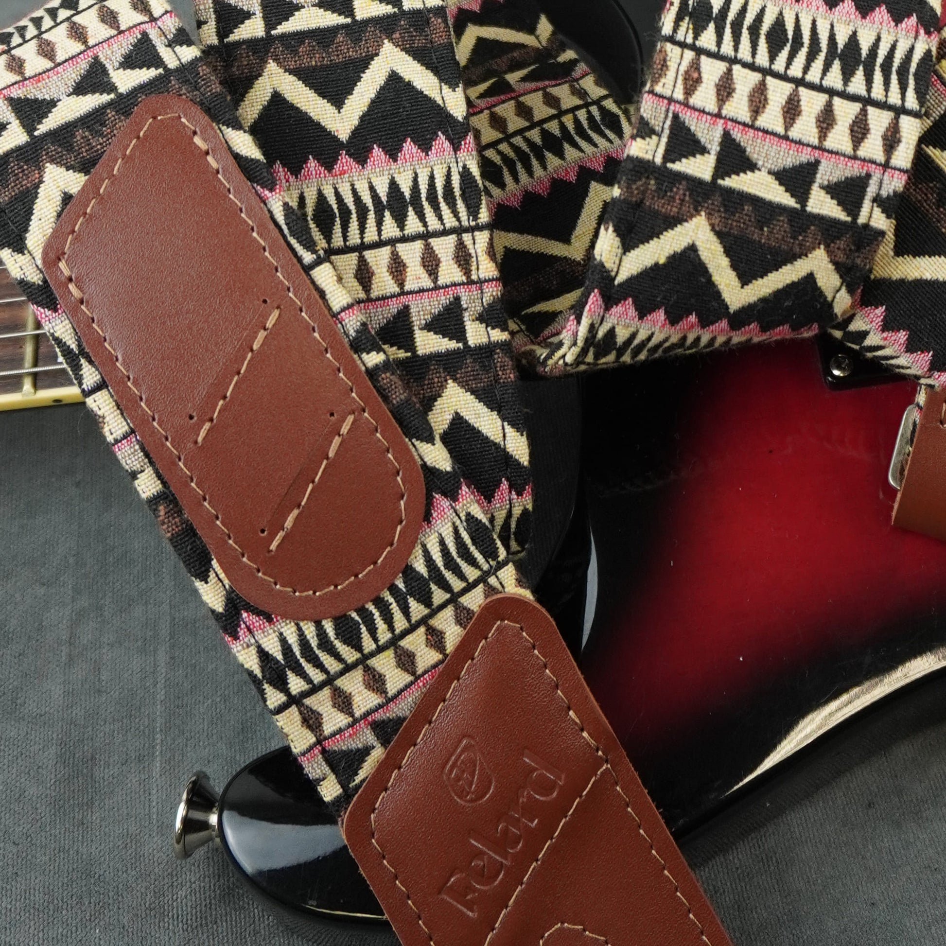 A close-up of a guitar strap with a geometric patterned fabric and brown leather patches, shown resting on a red electric guitar. The leather patch is embossed with the brand name 'Poldri'.