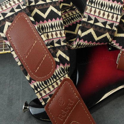 A close-up of a guitar strap with a geometric patterned fabric and brown leather patches, shown resting on a red electric guitar. The leather patch is embossed with the brand name 'Poldri'.