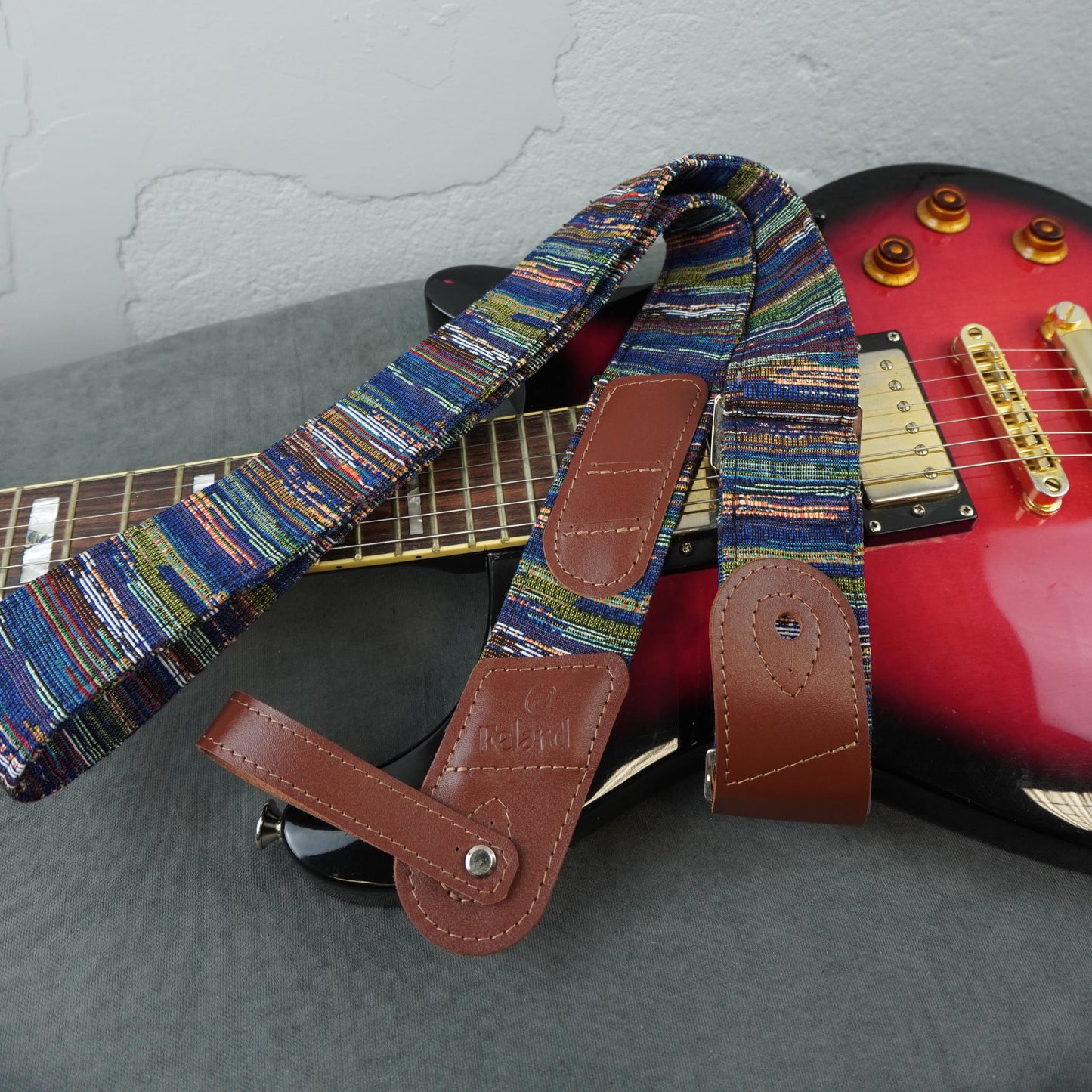 A close-up of a guitar strap with a multicolored woven pattern and brown leather ends, draped over a red electric guitar on a gray surface against a light wall.