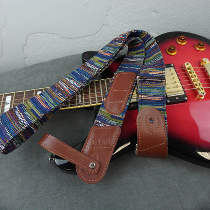 A close-up of a guitar strap with a multicolored woven pattern and brown leather ends, draped over a red electric guitar on a gray surface against a light wall.