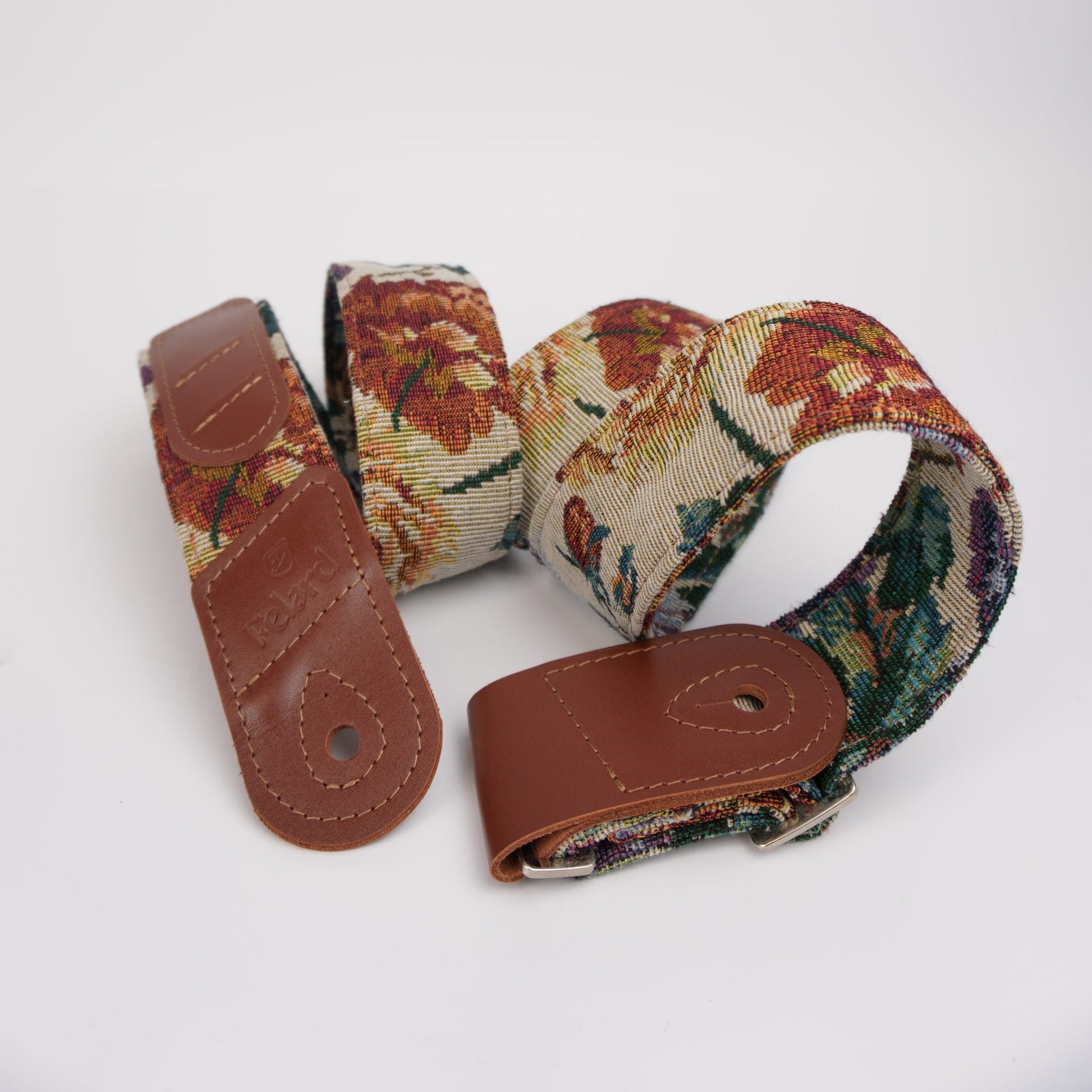 A woven guitar strap with a floral tapestry pattern in red, green, and beige, featuring brown leather end caps and a silver metal buckle, displayed on a clean white background.
