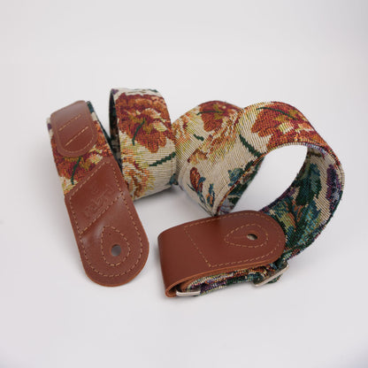 A woven guitar strap with a floral tapestry pattern in red, green, and beige, featuring brown leather end caps and a silver metal buckle, displayed on a clean white background.