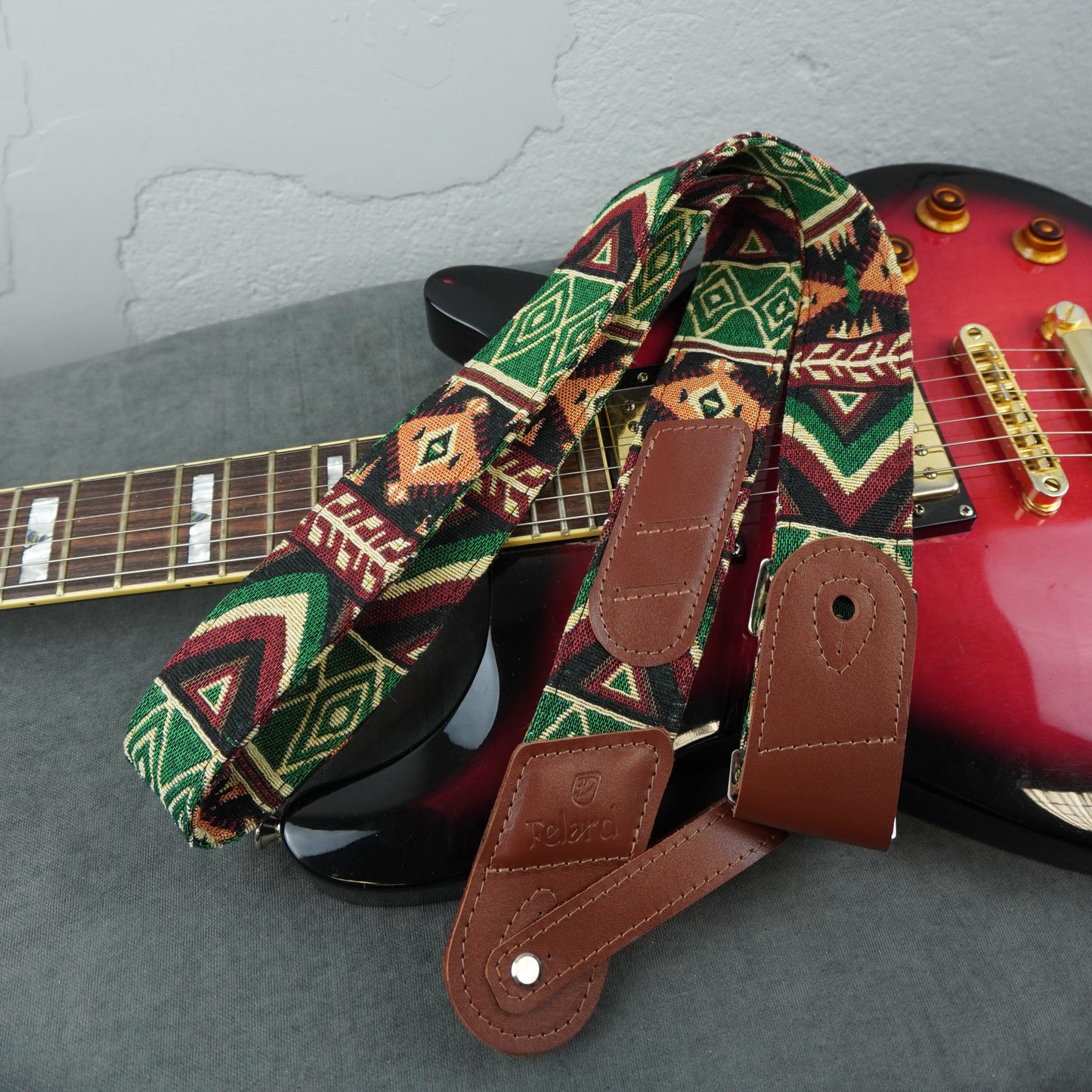 A guitar strap with a colorful geometric tribal pattern in green, red, orange, and black, combined with brown leather ends, draped over a red electric guitar on a gray surface.