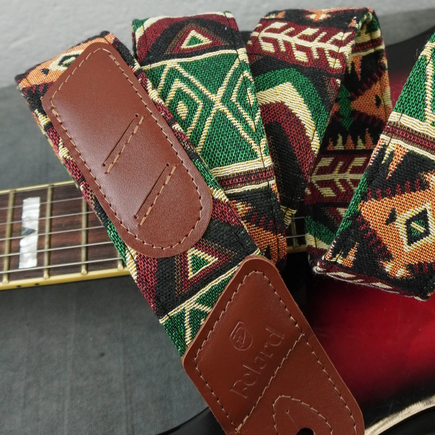 A close-up of a guitar strap with a colorful geometric woven pattern in green, red, black, and beige, featuring brown leather end pieces. A portion of a guitar neck is visible in the background.