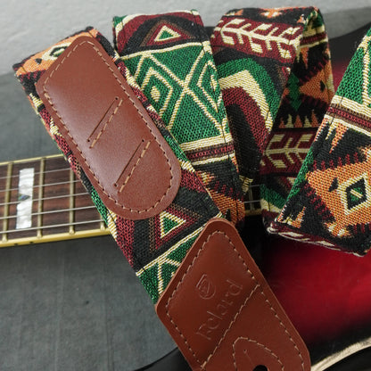 A close-up of a guitar strap with a colorful geometric woven pattern in green, red, black, and beige, featuring brown leather end pieces. A portion of a guitar neck is visible in the background.