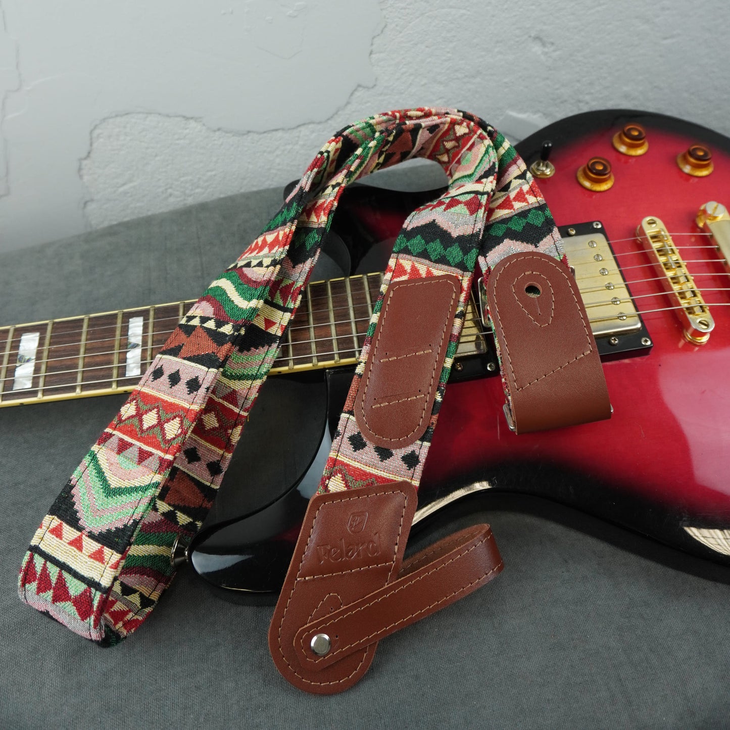 A multicolor geometric patterned electric guitar strap with brown leather ends, draped over a red and black electric guitar on a gray surface.