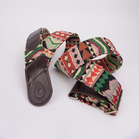 A woven fabric guitar strap with a Southwestern geometric kilim pattern in red, green, beige, and black, featuring dark brown leather ends.