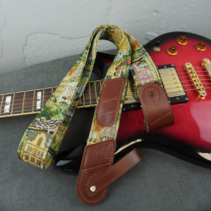 A guitar strap with a colorful cityscape print and brown leather ends, draped over a red electric guitar on a gray surface against a light gray wall.