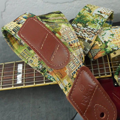 A close-up of a guitar strap with a multicolor cityscape needlepoint pattern and brown leather patches, shown resting on a guitar.