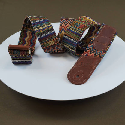 Guitar strap with a colorful geometric woven pattern and brown leather end pieces, displayed on a white round plate against a dark brown background.