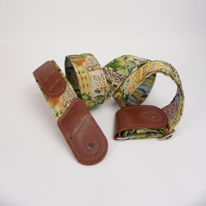 A canvas guitar strap featuring a colorful cityscape pattern with brown leather end caps, displayed on a clean white background.